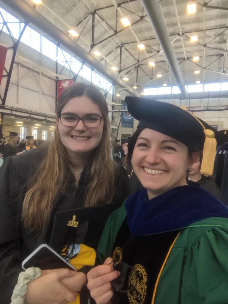 A graduate student and a faculty member, both in full graduation regalia, posing for a photo in a gymnasium.