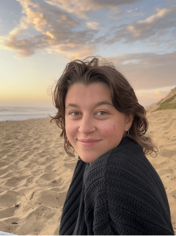 Portrait of Lola Holcomb, wearing a block sweater on a beach at sunset