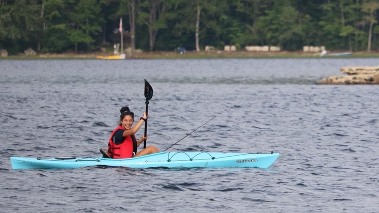 Louisa Colucci. Young woman with dark hair tied in a bun is sitting in a kayak with her paddle raised. She is wearing a black top and a red life-vest over it, in a light blue, single-person kayak. The kayak is in the water, most likely a lake, with the forested shoreline in the background.