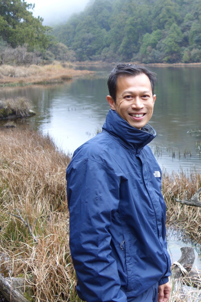 Dr. Sonny Lee is a blue rainjacket standing in front of a forested river on a cloudy day.